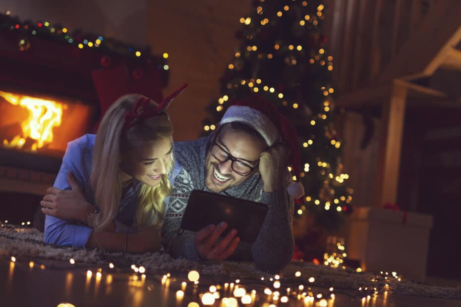 A man and woman relaxing on the floor in front of a Christmas tree, captivated by their latest tech gadgets.